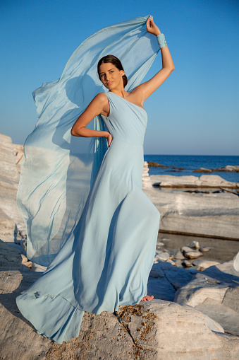 Beautiful young fashion model with brown hair back wearing long light blue dress, standing on the rocky coastline with hand on hip and lifting silky gown, beauty and fashion industry