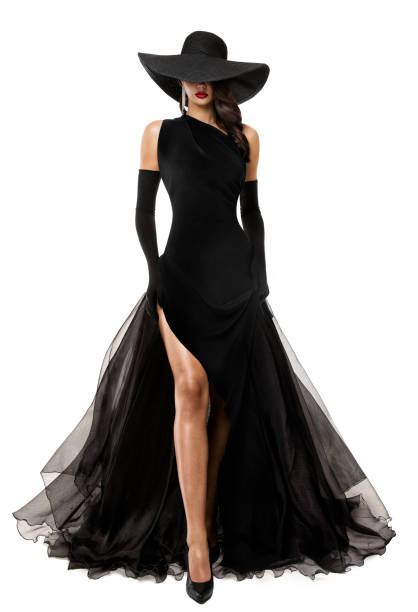 Elegant Lady in Long Black Dress and Hat. Fashion Woman in evening Luxury Gown with Slit over White. Mysterious Beautiful Girl showing Sexy Leg in Flying Skirt stock photo