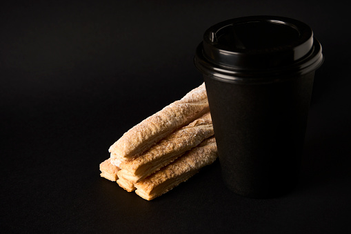 Paper coffee cup and Puff pastry sticks with cinnamon on black background with copy space. Take away coffee and breadsticks sprinkled with sugar, close-up