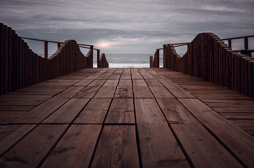 Long wooden pier boardwalk in Quiaios beach with low viewpoint perspective leading to the Atlantic ocean and cloudy autumn sunset sky. Leading lines with selective focus and empty space for text