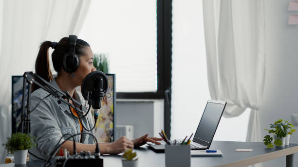 istock Internet radio show host talking on microphone while checking subject list on laptop. 1441371729