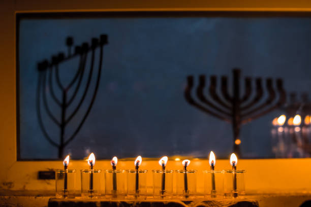 Glass vials hold oil and burning wicks in a menorah glowing brightly beneath a window with unlit menorahs on the eighth night of the Jewish festival of Hanukkah.