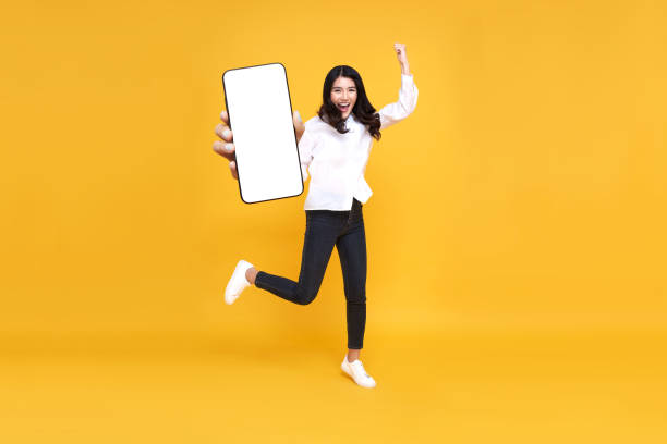 istock Happy young asian woman showing empty mobile phone screen while jumping up on yellow studio background. full body length. Excited lady recommending nice and useful mobile application 1441380364