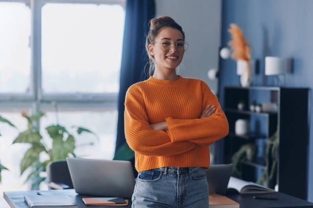 Confident woman keeping arms crosses and smiling while standing near her working place in office stock photo