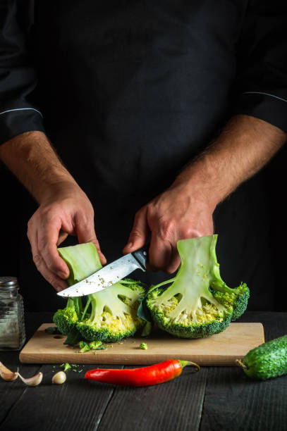 istock Cook cuts fresh Broccoli with a knife for salad on vintage kitchen table with fresh vegetables. Cooking and restaurant or cafe concept 1441383046