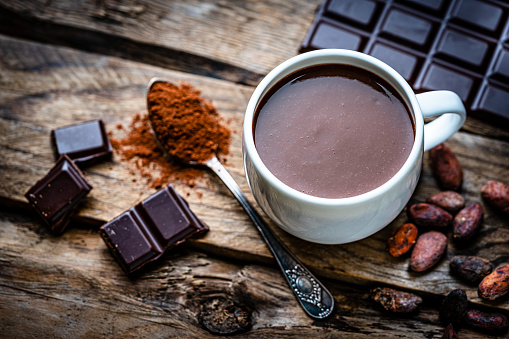 Sweet food: high angle view of a hot chocolate mug shot on rustic wooden table. Cocoa beans, dark chocolate bar and pieces and a spoon filled with chocolate powder complete the composition. High resolution 42Mp studio digital capture taken with Sony A7rII and Sony FE 90mm f2.8 macro G OSS lens