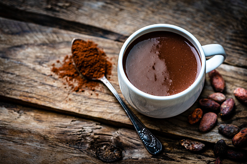 Sweet food: overhead view of a hot chocolate mug shot on rustic wooden table. Cocoa beans and a spoon filled with chocolate powder complete the composition. High resolution 42Mp studio digital capture taken with Sony A7rII and Sony FE 90mm f2.8 macro G OSS lens