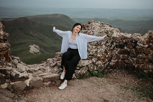 Happy woman tourist hiking up mountain enjoying nature. Landscape in cloudy gloomy windy weather. Female in white shirt with open arms outstretched in joy, enjoying travel. Freedom concept