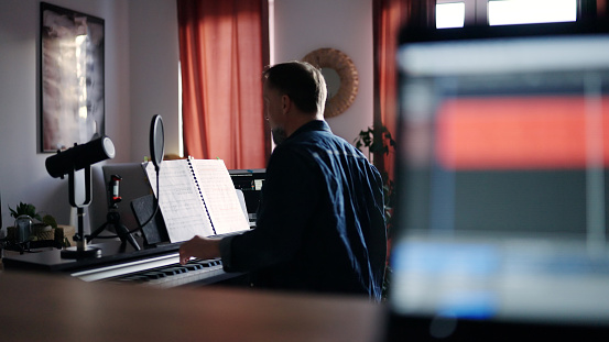 Rear view of man seated playing piano and recording musical composition in a laptop. Focus on background, blurred foreground.