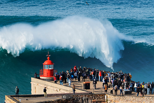 People watching the big giant waves crashing near the Fort of Nazare Lighthouse in Nazare, Portugal. Biggest waves in the world. Touristic destination for surfing. Amazing destinations. Nazare Canyon