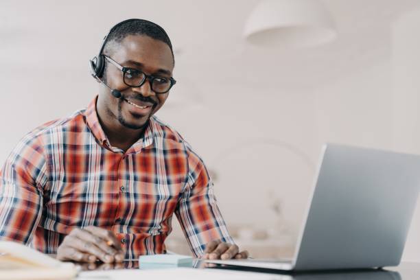 istock Smiling african american man in wireless headset sits at desk, studying online on laptop. E-learning 1441391692