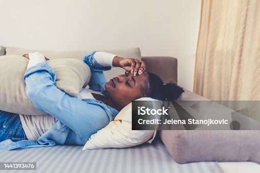 istock Cropped short of a woman holding a bridge pillow against her stomach on the sofa at home. Photo of a young woman having stomach pain while lying on the couch at home. 1441393476