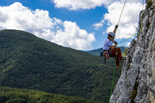 Climbing scene of a guy on a rock wall with a green forest and a light blue cloudy sky behind him