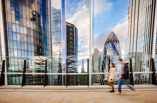 Color image depicting blurred motion of two people going about their business set against the backdrop of the modern futuristic London skyline, including the distinctive architecture of the Gherkin (30 St Mary Axe) building.