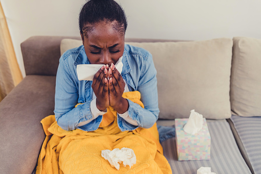 Shot of a young woman blowing her nose with a tissue at home. Young woman with a cold, sitting on the couch, sneezing into a tissue.