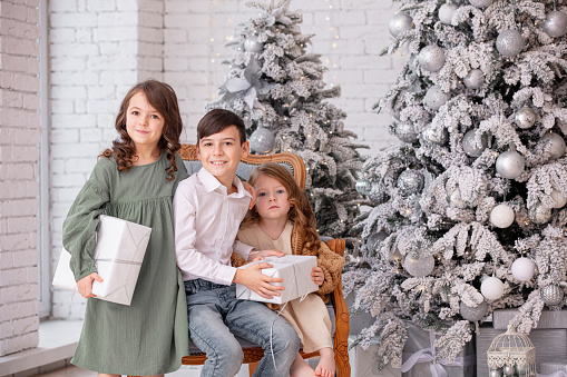 Three children, two girls and a boy, happy together at home celebrating Christmas at a decorated Christmas tree with gifts in their hands