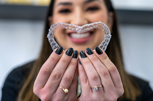 Woman smiling and holding Clear Aligners/ Invisible braces forming a heart shape. Woman with perfect smile after invisible aligners treatment.