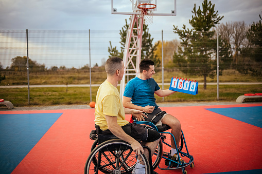 Two wheelchair-bound men on outdoor basketball court.