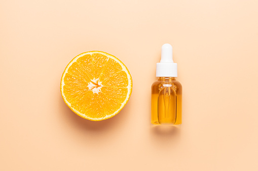 Glass bottle with pipette and half orange on beige background, top view