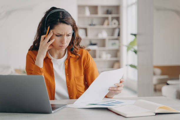 istock Focused professional woman wearing headset reading document, working at laptop at home. Remote work 1441403736