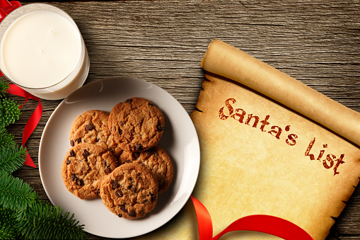 Santa's list rests underneath a plate of cookies and a glass of milk in a scene that is framed  by a red ribbon and a bough from a Christmas tree.