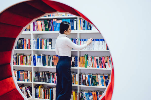 Side view of focused Asian female student in casual outfit standing in front of bookcase in university library searching for book