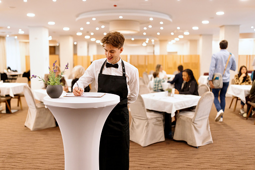 Waiter looks at the guest checklist and greets the hotel guests during breakfast