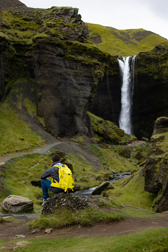One unrecognizable tourist enjoying the nature's wonders on Iceland, surrounded by grassy and rocky terrain, observing the waterfall in the distance