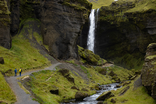 Unrecognizable tourists walking around the Iceland's beautiful nature, surrounded by high rocky cliffs and a tall waterfall