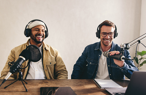 Male radio presenters having a great time on a live show. Two men smiling happily while recording an audio broadcast in a studio. College content creators co-hosting an internet podcast.