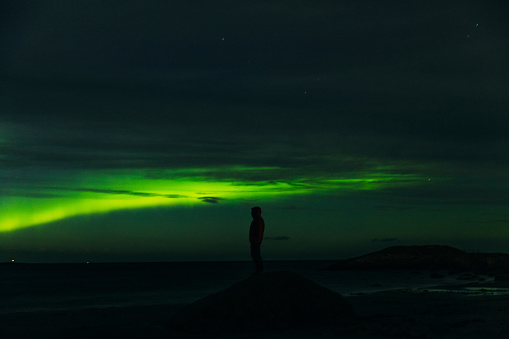 Silhouette of a male staying on the stone at the beach looking at the bright Aurora borealis in the sky during the autumn night in More og Romsdal