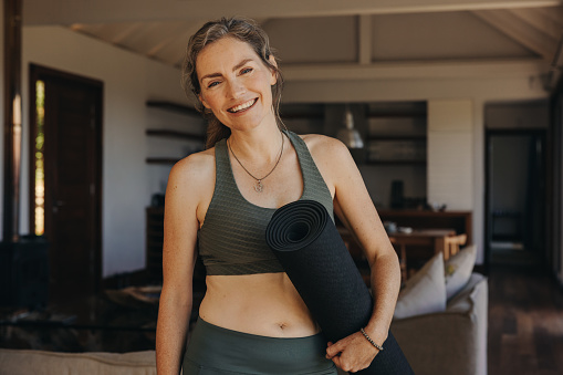 Happy senior woman smiling at the camera while holding an exercise mat. Cheerful elderly woman preparing to start a yoga session at home. Mature woman practicing a healthy workout routine.
