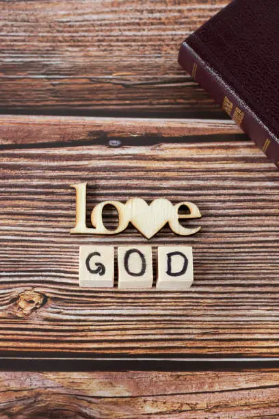Love God text written with wooden cubes and letters with heart shape on rustic background with closed Holy Bible Book. Vertical shot. Top view. Christian biblical concept.