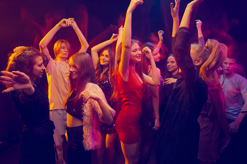 Group of young people attending party, dancing, having fun over dark background in neon light. Friends gathering together. Concept of youth culture, leisure time activity, fun, lifestyle
