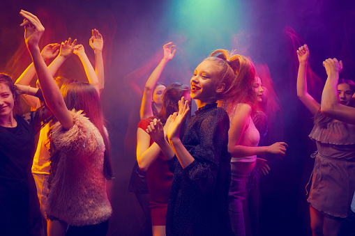 Group of young people, girls attending party, dancing, having fun over dark background in neon with mixed light. Concept of youth culture, leisure time activity, fun, lifestyle, celebration