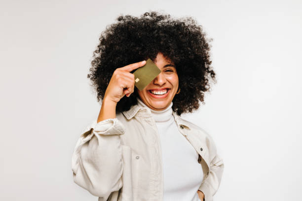 Happy young woman holding a credit card over her eye Happy young woman smiling at the camera while holding a credit card over her eye. Cheerful woman with curly hair recommending electronic banking and online shopping. credit card stock pictures, royalty-free photos & images