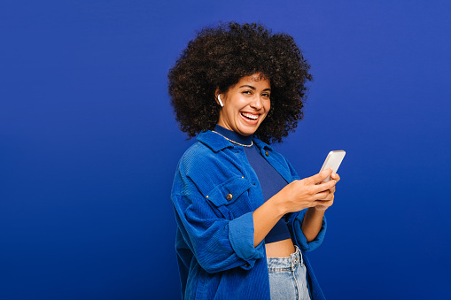 Cheerful woman smiling at the camera while playing music using a smartphone and earbuds. Young woman with curly hair enjoying her favourite playlist while standing against a blue background.