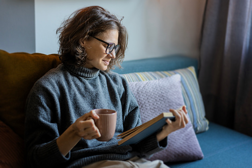 Young smiling cheerful woman in a warm sweater reading a book while sitting on the sofa with a mug of coffee in her hand