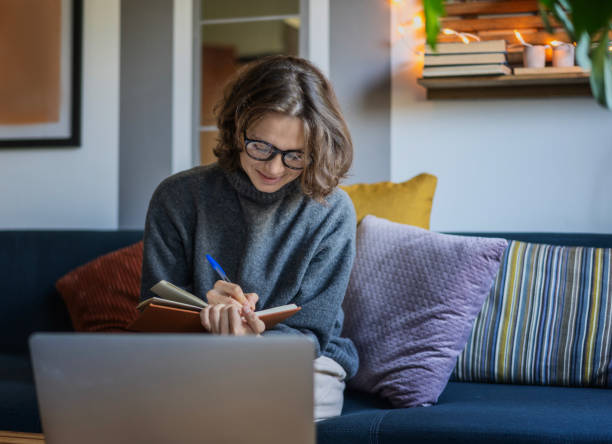 Young woman in a warm sweater sitting on a sofa with a laptop making notes in a notebook, work and education online concept