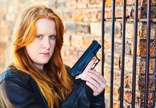 Young woman with flaming red hair with a semi-automatic pistol by the window of a house, protected by security bars.