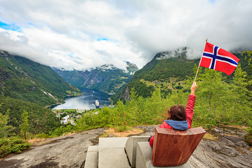 Travel adventure concept. Female tourist enjoying view over Geirangerfjord from Flydalsjuvet viewpoint, holding norwegian flag.