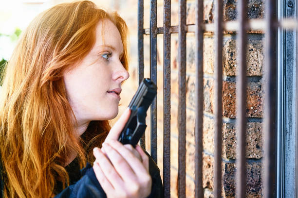 Young woman with flaming red hair with a semi-automatic pistol by the window of a house, protected by security bars.