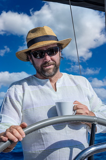A man in a straw hat drinks coffee while driving a sailboat.