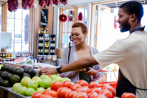 The mid adult customer smiles as the young adult male clerk helps her with the produce at the small market.