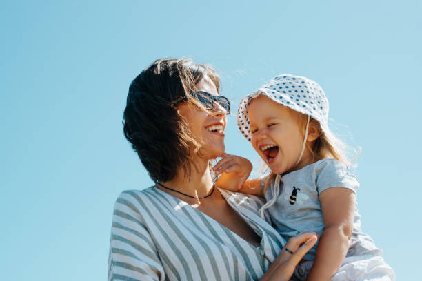 Positive family portrait laughing mom and daughter outdoors. Cheerful young beautiful mother holding laughing baby in her arms against blue sky stock photo