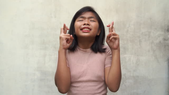 Asian teenage girl wishing for good luck, finger crossed for pray , white old wall background