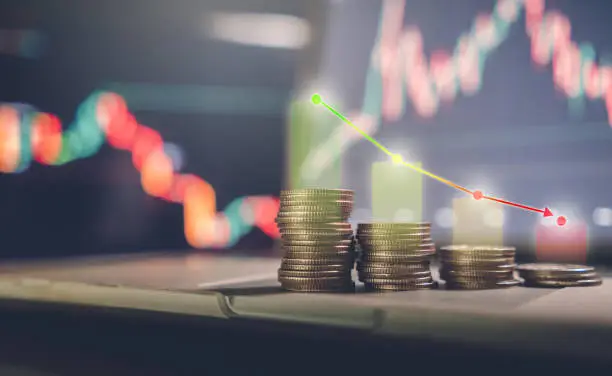 Photo of investment concept, stacked coins with a background monitor showing data graphs with an arrow pointing to a low point.