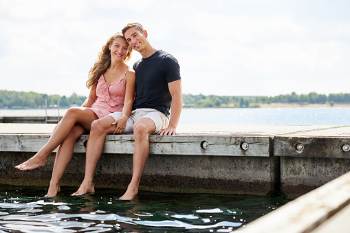 a young couple sits on a jetty by a lake in germany and smiles at the camera