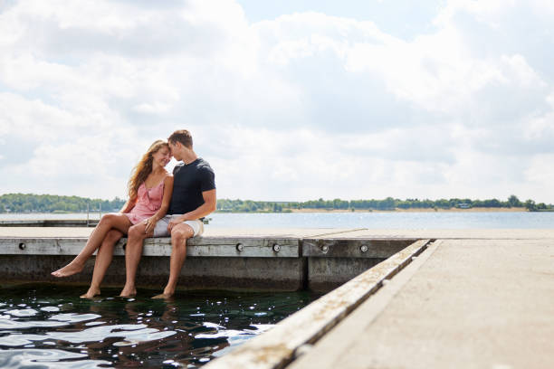 a young couple sits on a jetty at a lake in germany and snuggles their heads together