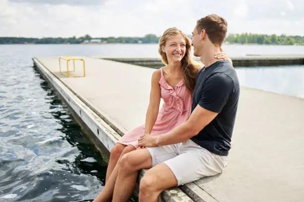 a young couple sits on a jetty by a lake in germany and smiles at teach other